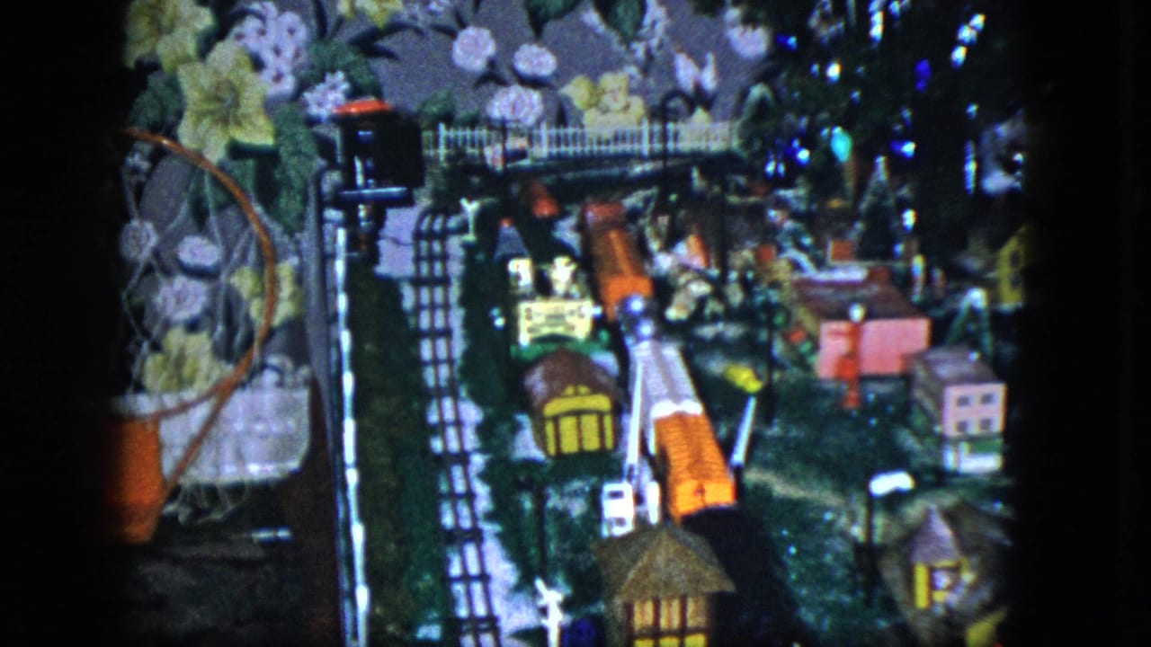 1951 Cleveland home-movie frame showing a lit model railroad scene with tracks, miniature houses, and a floral wallpaper backdrop.