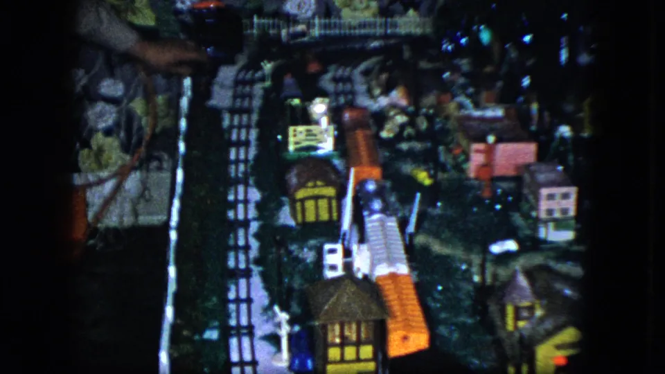 1951 Cleveland home-movie frame with a hand adjusting a model train display beside tracks, small houses, and lights.