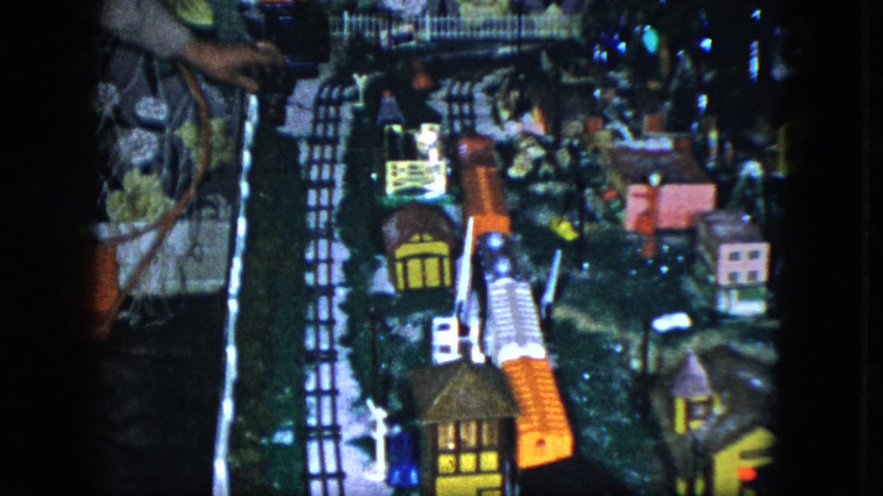 Model train layout with tracks, miniature houses, lights, and a hand at the left edge.