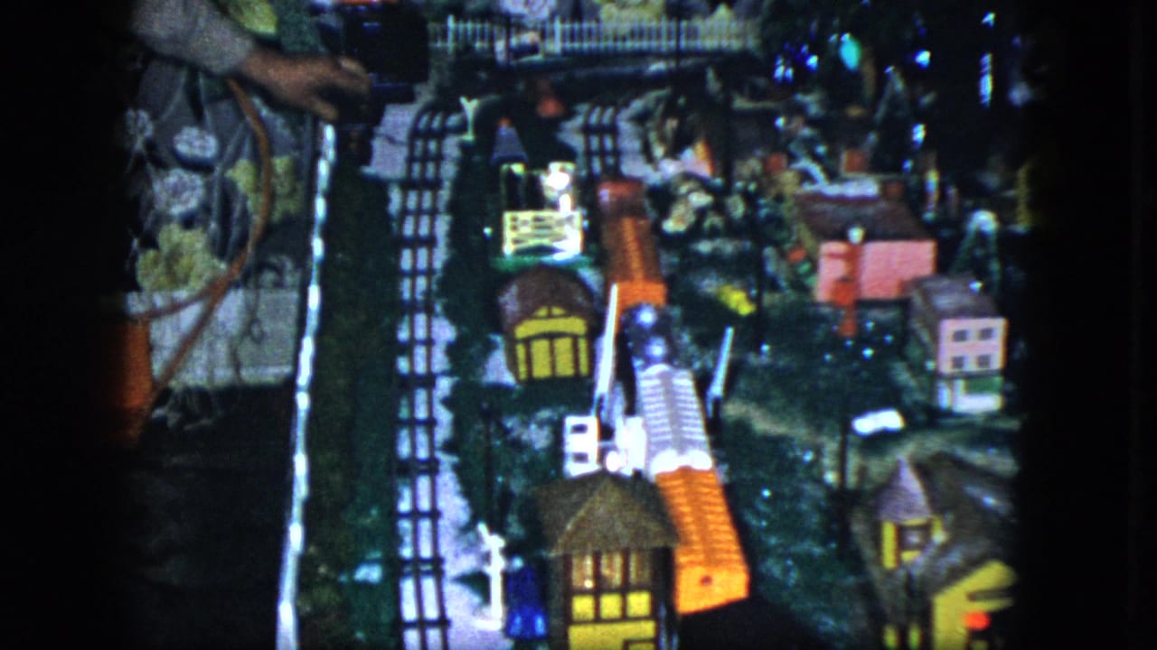 1951 Cleveland home-movie frame of a model railroad layout, with illuminated buildings, tiny houses, tracks, fence, and orange train cars.