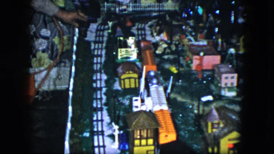 1951 Cleveland 8mm frame showing a miniature railroad scene with lit houses, tracks, trees, a small fence, and a hand near the edge.