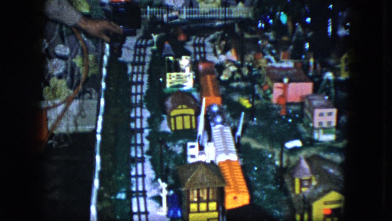 1951 Cleveland home-movie frame showing a tabletop electric train village with tracks, small houses, trees, and a partial adult figure nearby.