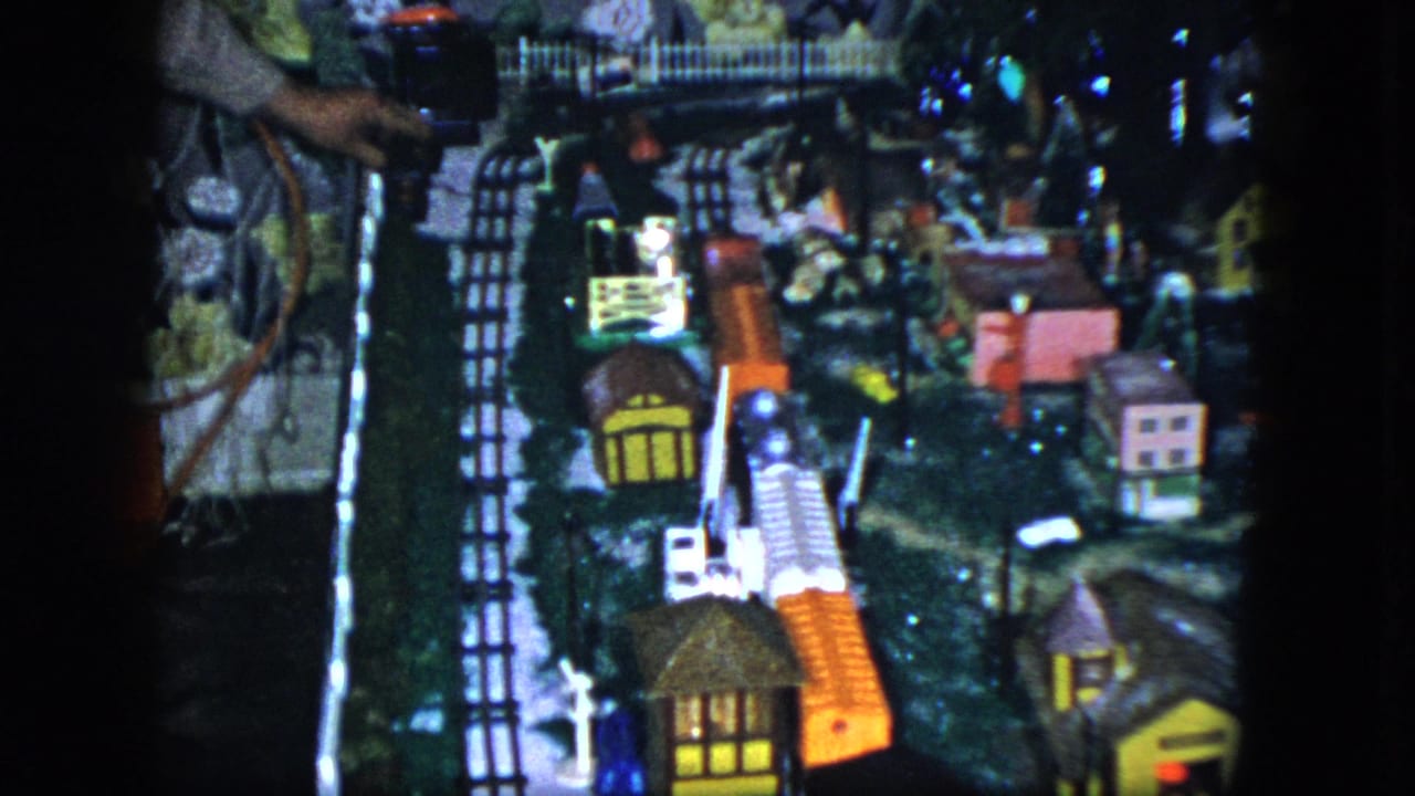 1951 Cleveland 8mm frame of a tabletop train village, with curved track, colorful model buildings, trees, and shadowed display edges.