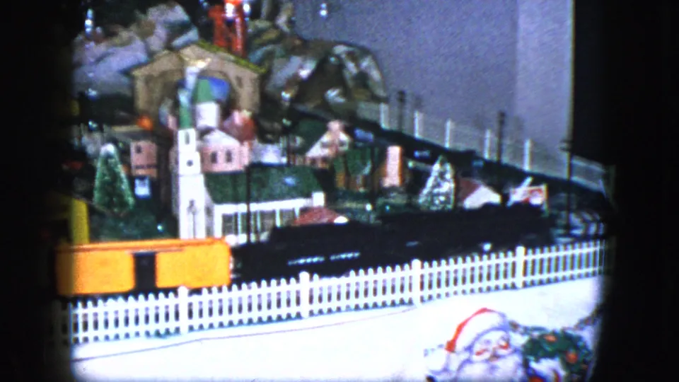 1951 Cleveland home-movie frame showing a toy train scene with a white fence, miniature buildings, bridge, trees, and orange cars.