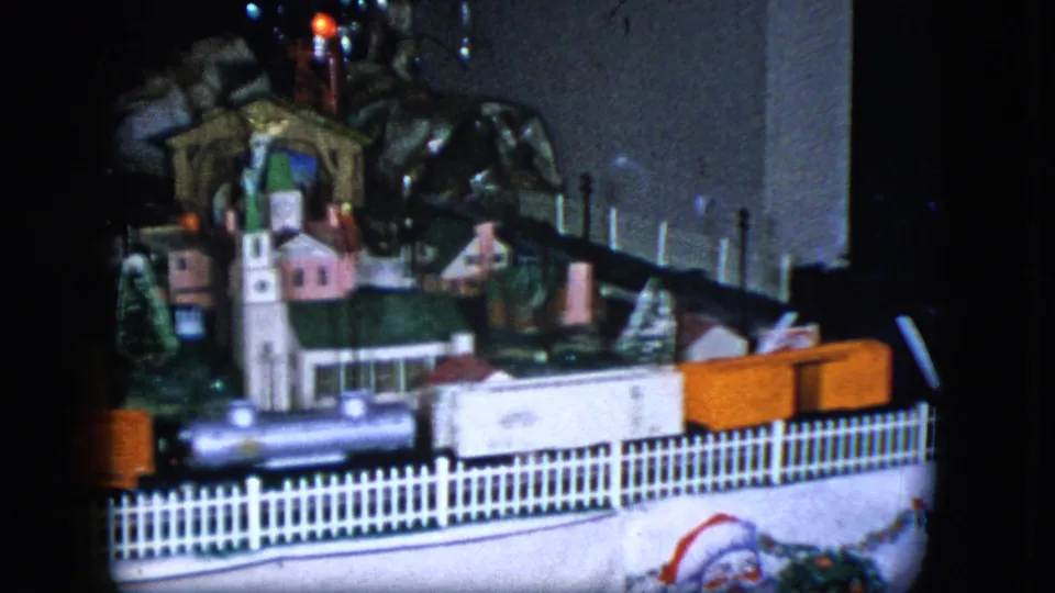 1951 Cleveland 8mm frame of a tabletop railroad set with train cars, white fence, village buildings, trees, bridge, and scenic rocks.