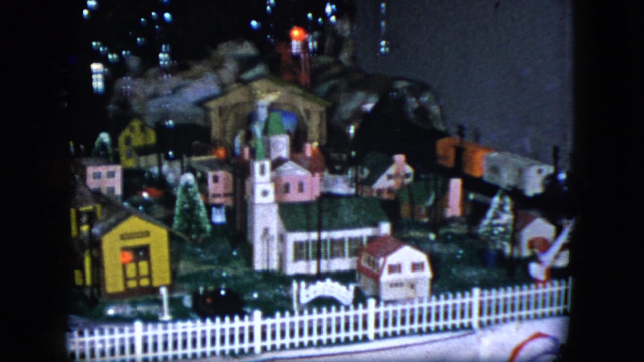 1951 Cleveland frame of a tabletop miniature village with a church, small houses, white fence, and toy train layout under colored lights.