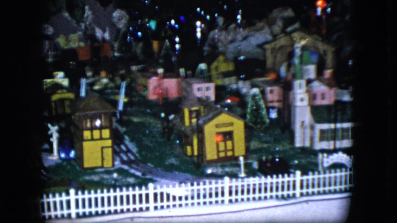 1951 Cleveland 8mm frame showing a miniature model train layout with houses, tracks, a white picket fence, and dark backdrop.