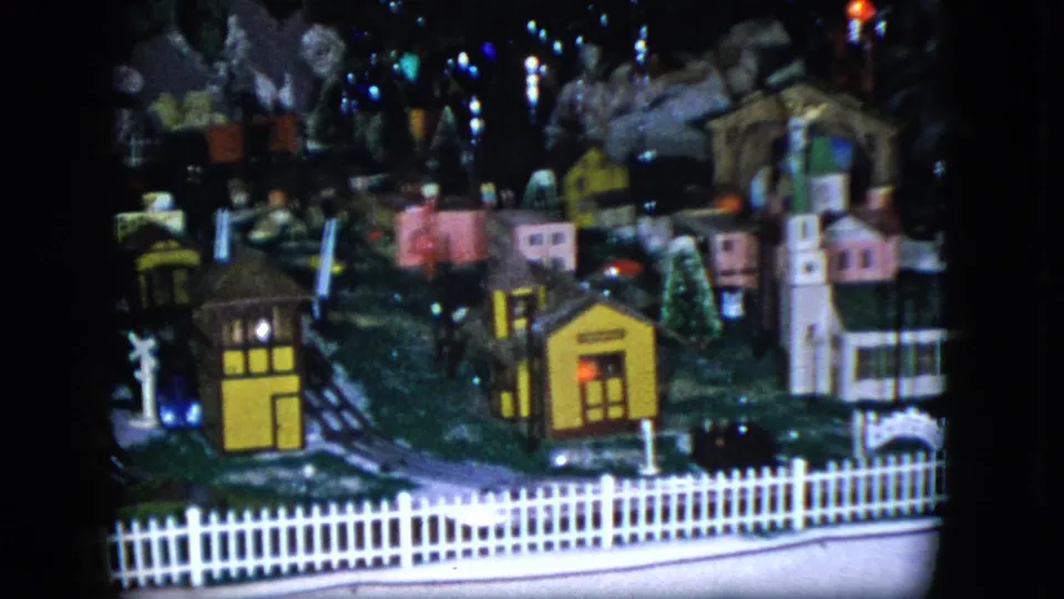 1951 Cleveland 8mm frame showing a miniature model train layout with houses, tracks, a white picket fence, and dark backdrop.