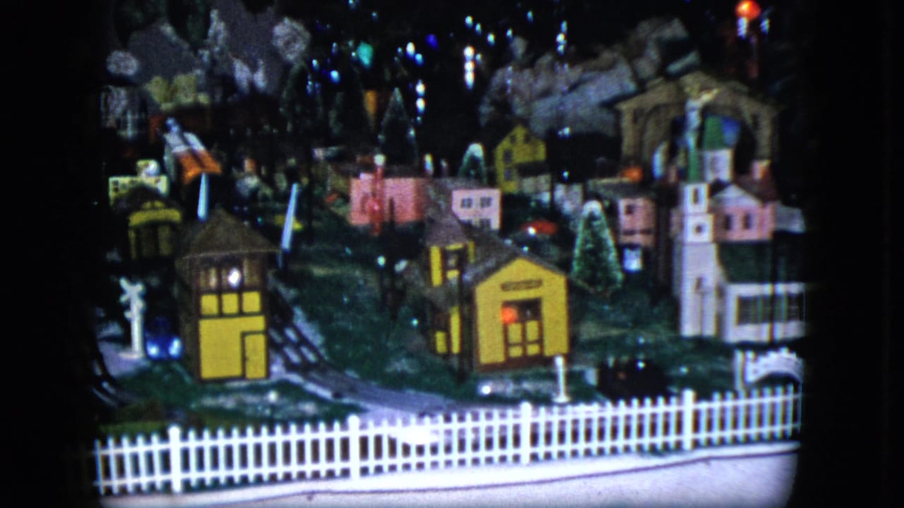 Model train layout with yellow building, pink houses, tracks, fence, trees, and dark background.