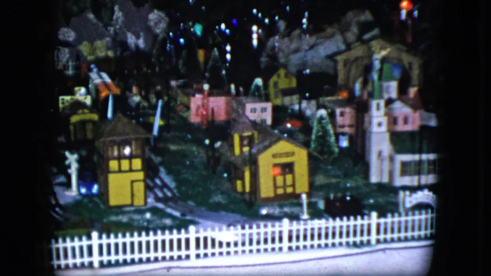 1951 Cleveland home-movie frame of a model train set, with a yellow building, small houses, tracks, fence, and trees.