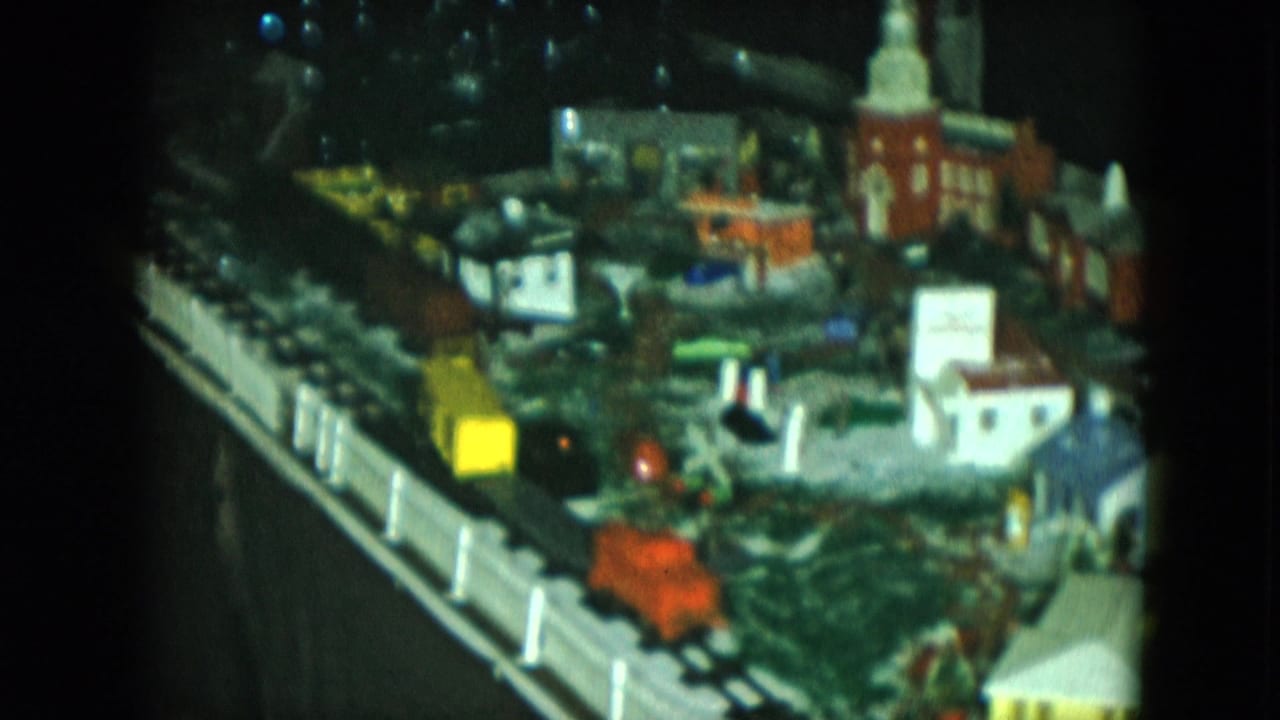 Model train layout with lit miniature town, church steeple, tracks, and white fencing.