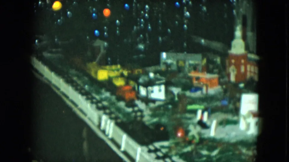Soft 1951 Cleveland home-movie frame showing a lit model train layout with tracks, small structures, fencing, and holiday-like bulbs.