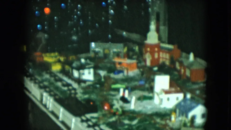 1951 Cleveland frame of a miniature railroad town display, with a church steeple, white fencing, small houses, and glowing colored lights.