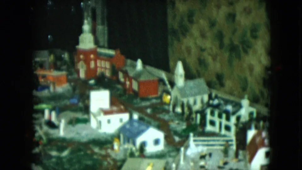 1951 Cleveland 8mm still showing a model train-set town with a church, small homes, track sections, and textured hillside scenery.