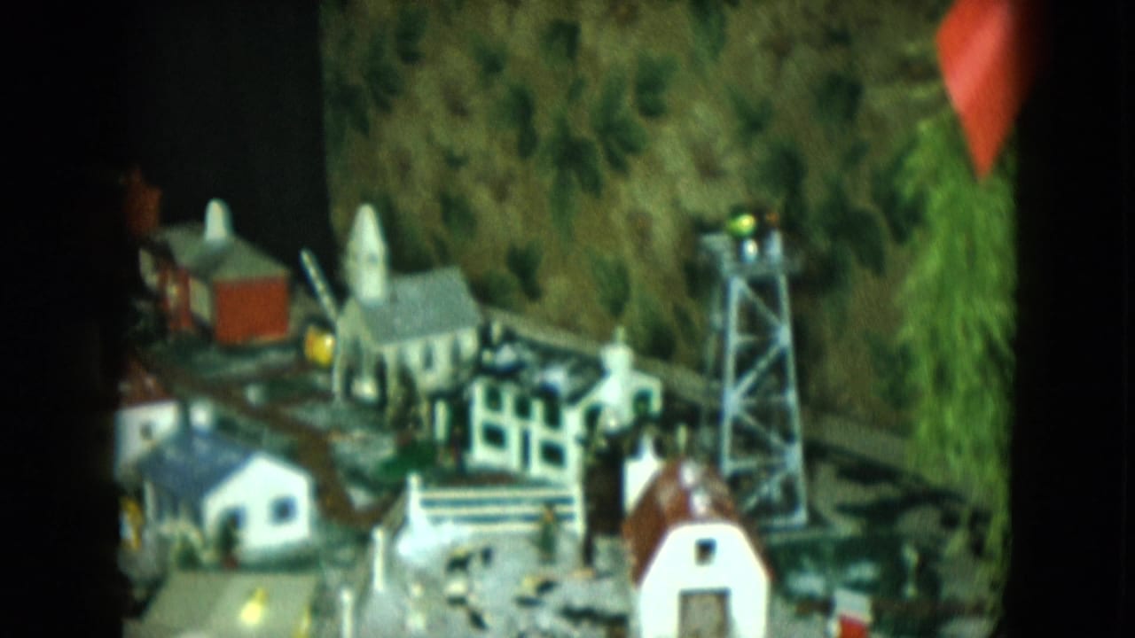 Model city scene with small buildings, tower, track, and painted foliage backdrop.