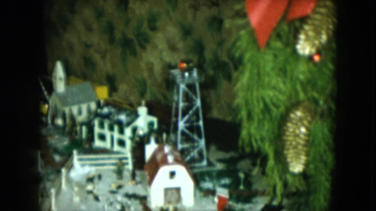 1951 Cleveland 8mm frame showing a Christmas model railroad town beside decorated evergreen branches, with a church, water tower, and small buildings.