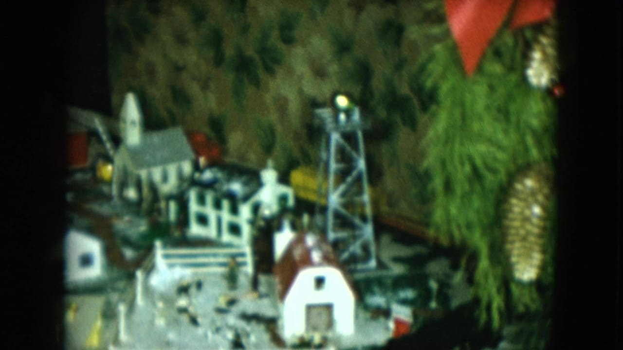 1951 Cleveland home-movie frame of a miniature railroad village with church steeple, tower, tracks, evergreen branches, pinecones, and red bow.