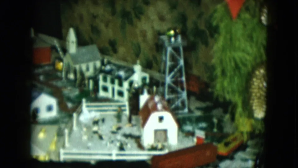 1951 Cleveland 8mm frame with the camera closer to the Christmas train set, showing a church, water tower, white buildings, tracks, and greenery.