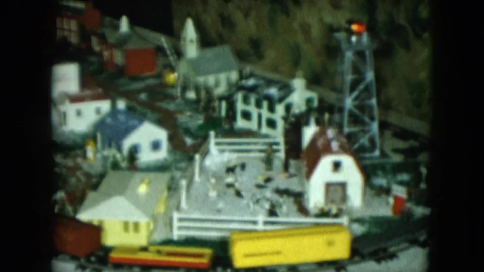 1951 Cleveland 8mm frame showing a wider Christmas train set with yellow rail car, miniature town buildings, church, water tower, and tree greenery.