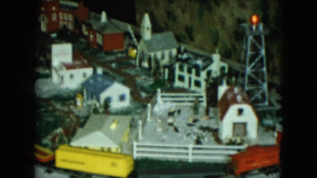1951 Cleveland 8mm frame showing a model train layout around a small snowy town with tiny buildings, fencing, and train cars.