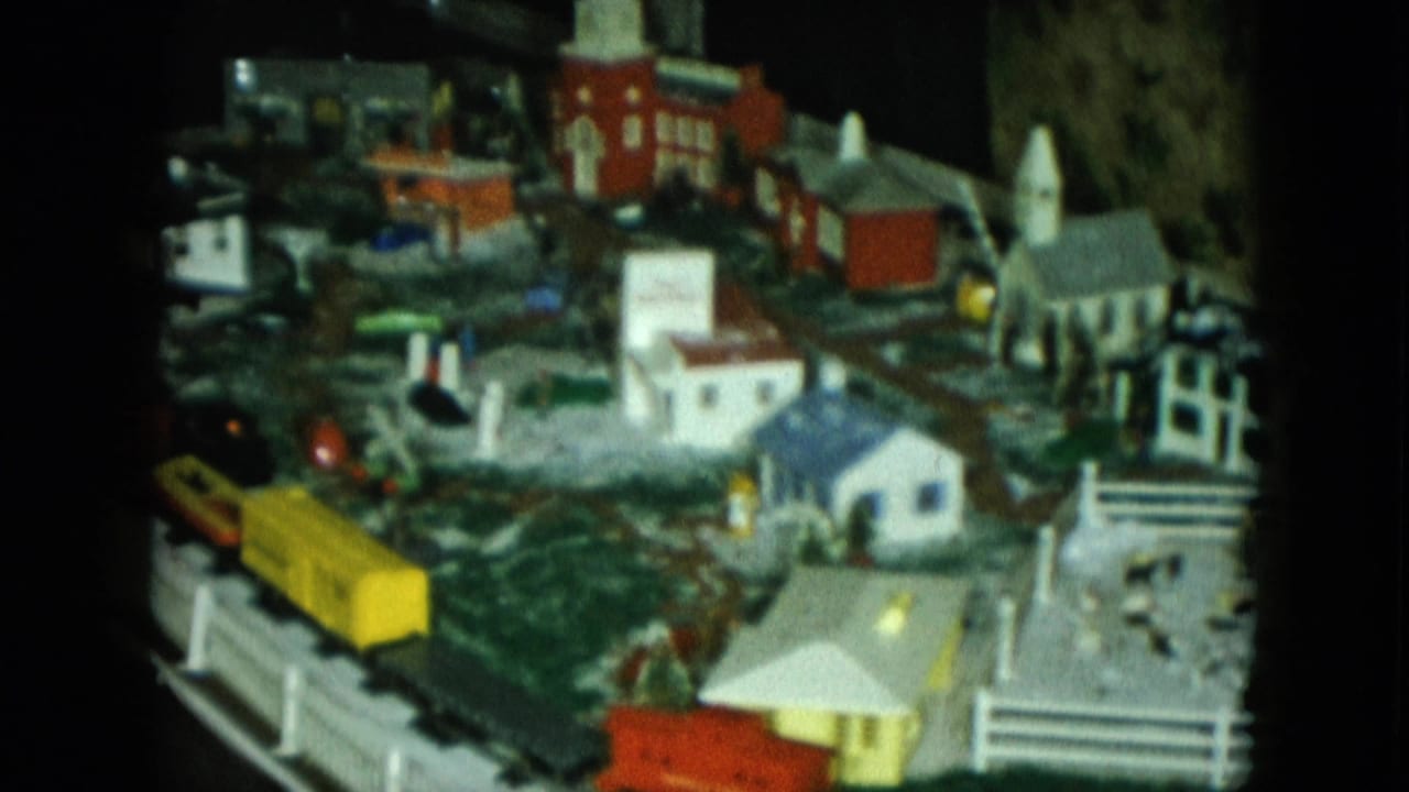 1951 Cleveland home-movie frame of a miniature railroad scene with yellow train car, snow-covered town details, and a church-like building.