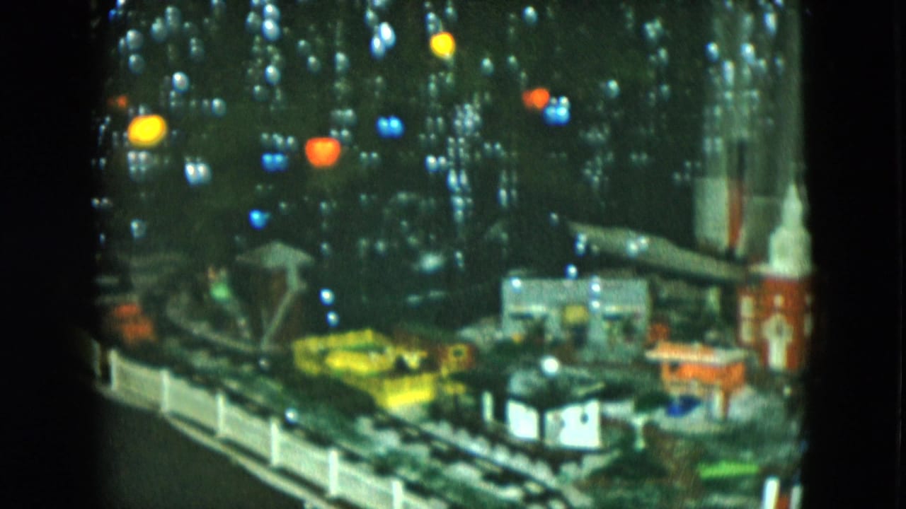 1951 Cleveland home-movie frame showing a model train scene with a red church, white steeple, small buildings, and Christmas lights.