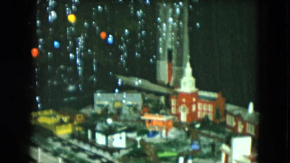 1951 8mm Cleveland holiday frame with a miniature red church, white steeple, nearby buildings, and train tracks under tree lights.