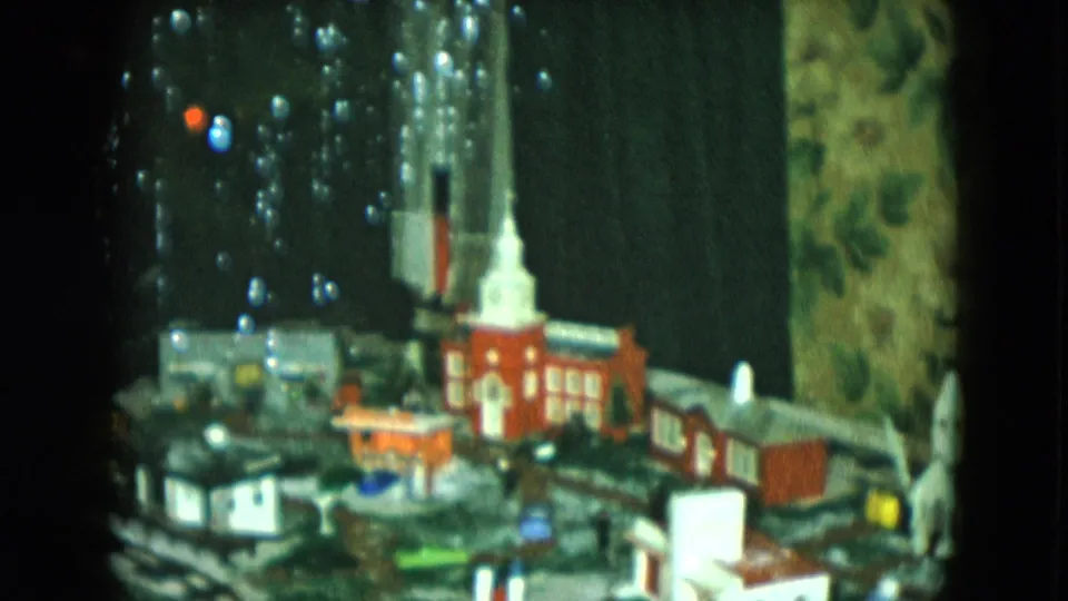 1951 Cleveland home-movie frame showing the Christmas village display with red church, rail layout, and the edge of the decorated tree.