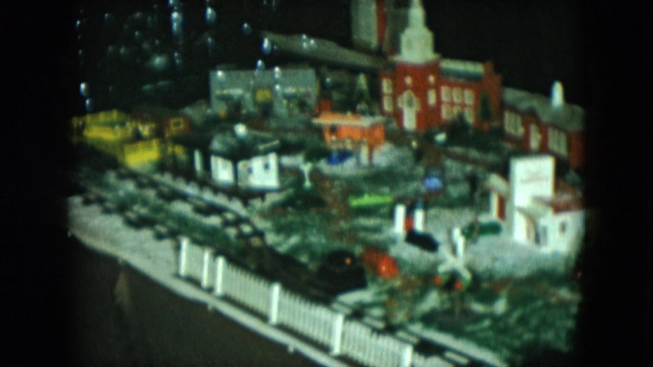 1951 Cleveland 8mm frame showing a black model locomotive moving through a miniature town display with buildings and fencing.