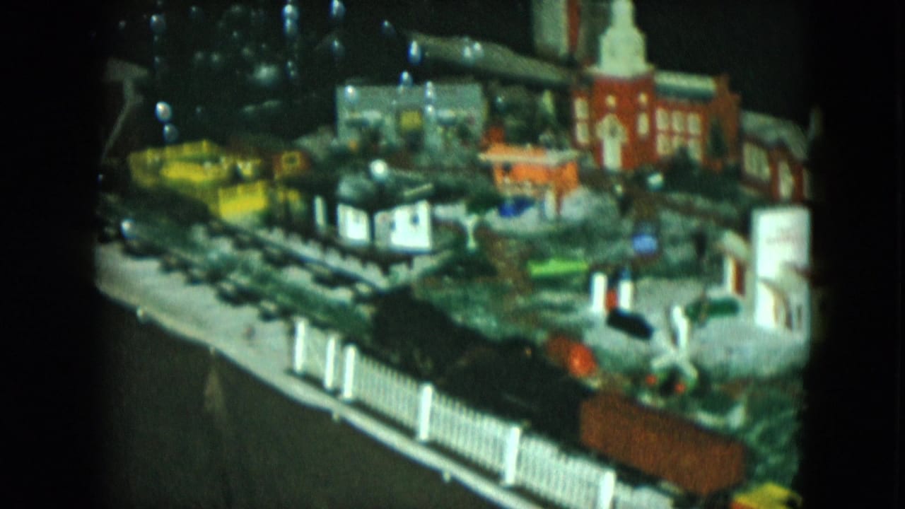 1951 Cleveland home-movie frame of a model train display with a black engine and orange freight car near a white fence.