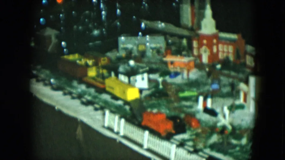 1951 Cleveland frame of a model train layout with yellow and orange cars passing a church-like miniature building and fence.