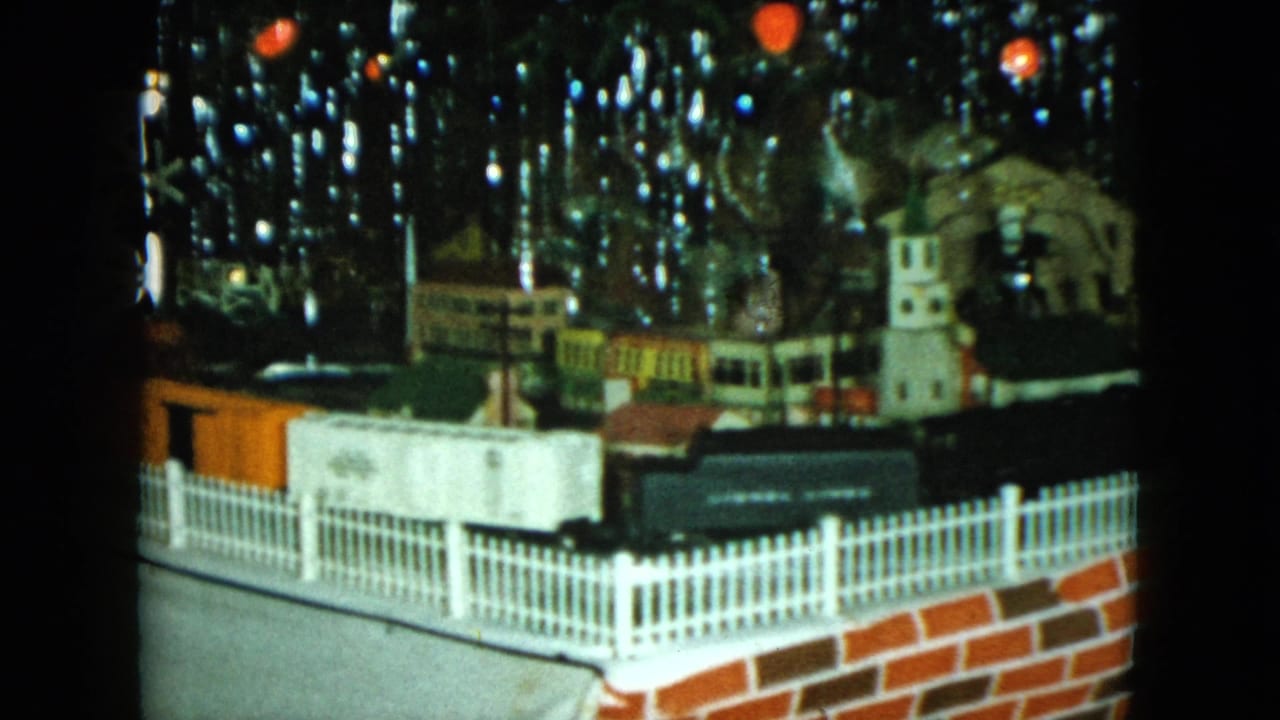 Black toy train passing a fenced miniature town with Christmas decorations behind it.