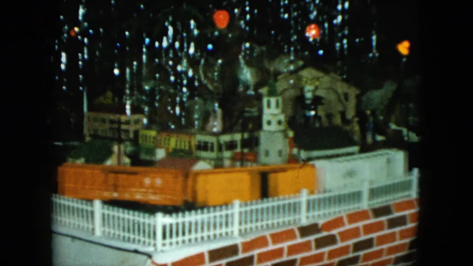 The toy train layout shows an orange railcar crossing the foreground of a Cleveland holiday miniature railroad scene.