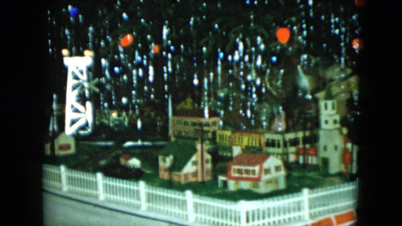 Miniature town with model houses, white fence, illuminated tower, and reflective holiday lights.