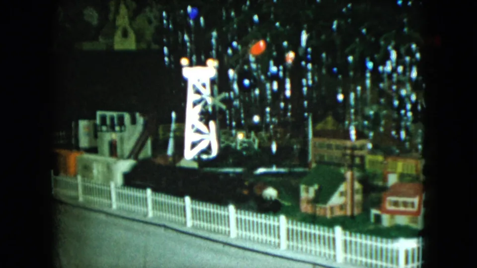 The 1951 8mm footage shows the miniature train display closer to the track and fence, with small buildings and bright reflections filling the background.