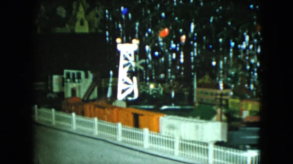 A small train passes through the 1951 Cleveland miniature town display, framed by a white fence, illuminated tower, model buildings, and reflected holiday lights.