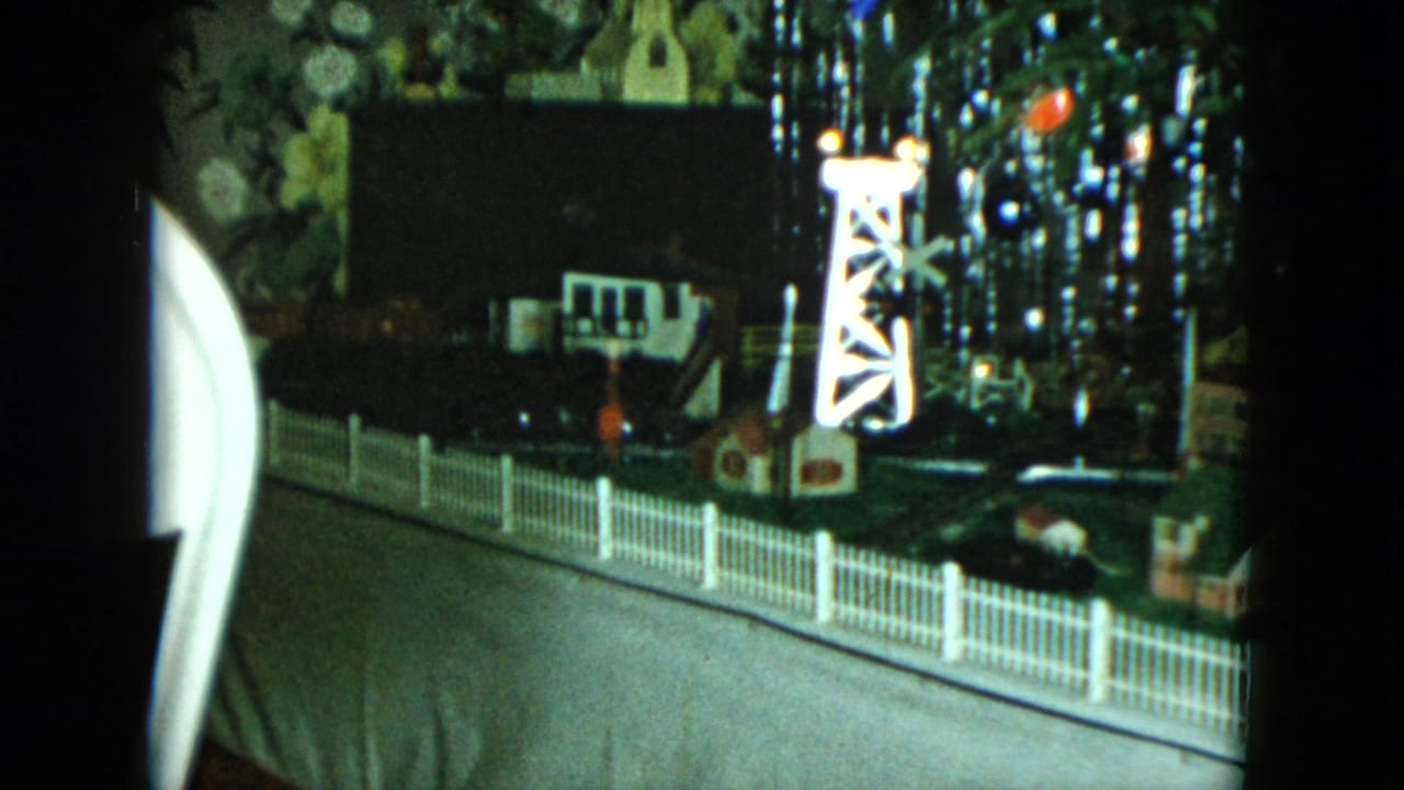 1951 Cleveland home-movie frame showing a tabletop model railroad scene with a white fence, lit tower, small buildings, and Christmas lights.