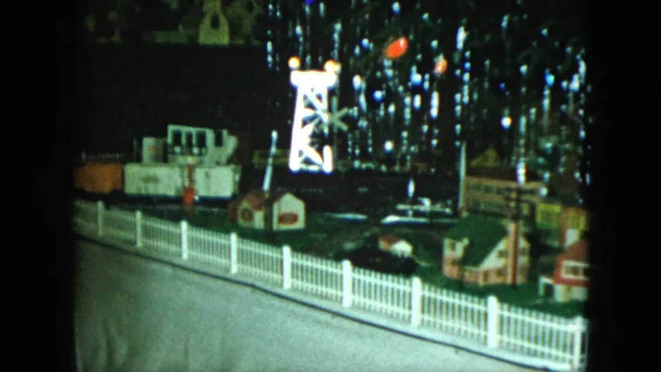 A closer 8mm frame shows the tabletop train scene in Cleveland, with fence sections, miniature structures, a bright tower, and holiday lights.