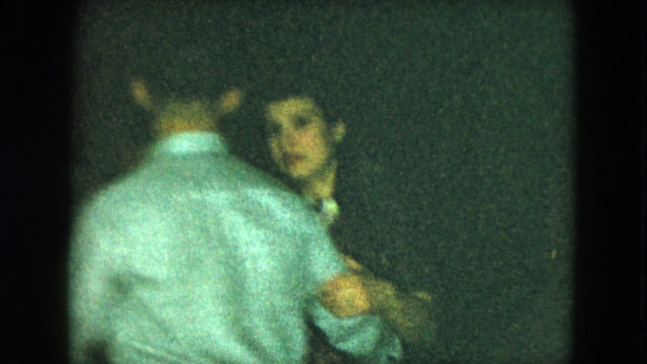 A dancing pair in light shirts, with one person looking toward the camera in a dark room.
