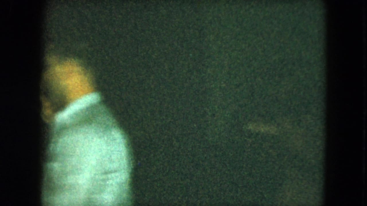 Man in a light collared shirt stands near the camera in a dim party scene.
