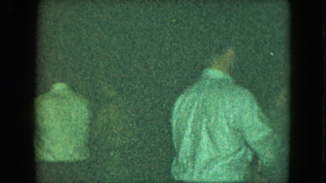 Two men in light shirts seen from behind in a dark, grainy room.
