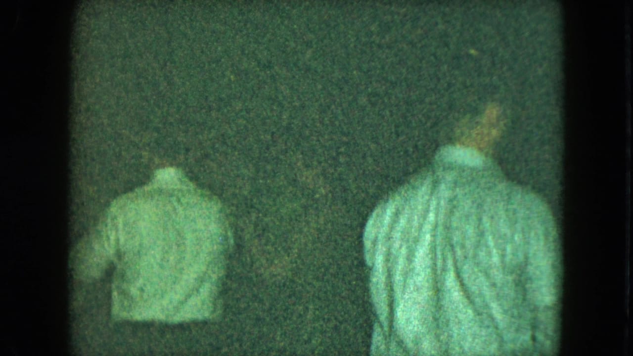 Two men from behind, one closer to the camera, in a dim green-tinted room.