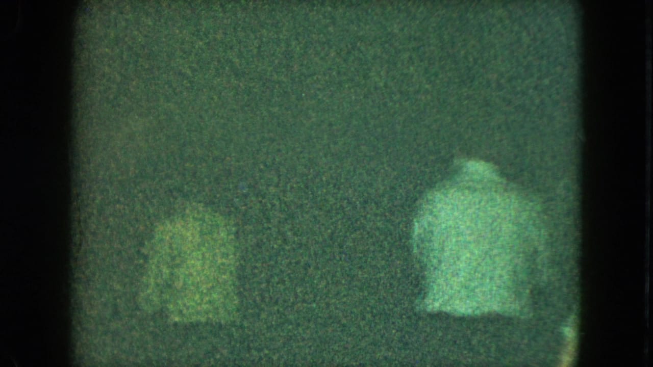 Dim green-tinted frame with blurry figures moving inside a dark room.