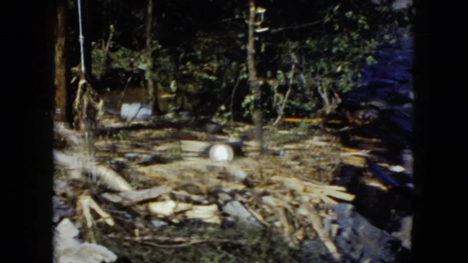 1951 Cleveland 8mm frame with a debris-covered wooded shoreline, broken logs, and turbulent water at the right edge.