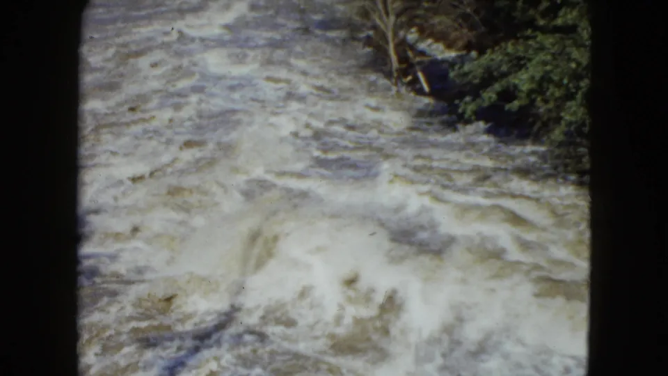 A vintage 8mm view captures foamy rapids below a wooded river edge, with soft color and home-movie gate borders.