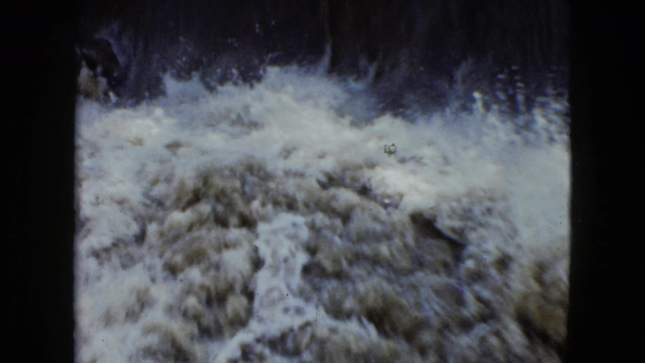 Foamy water churns below a dark small waterfall in a grainy 8mm frame.