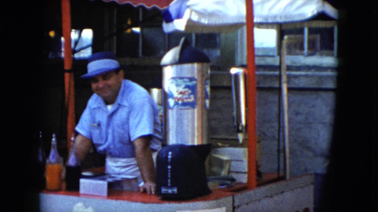 In the stated 1951 Cleveland footage, a smiling vendor stands behind a red-trimmed drink booth with bottles, a counter box, and a large dispenser.