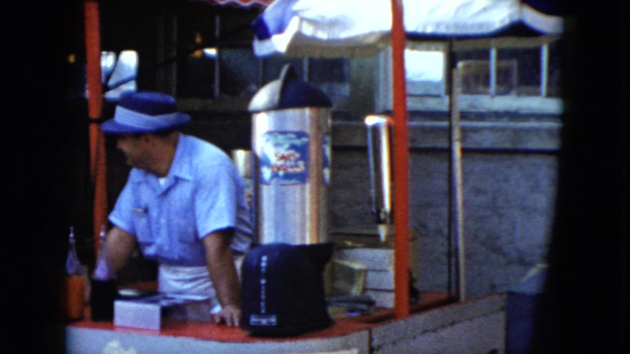 The vendor turns to his side at the Cleveland fairground-style booth, with bottled drinks, red posts, and a large drink dispenser visible.