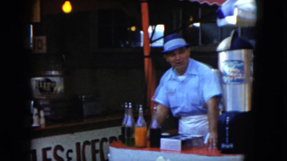 A blue-shirted vendor faces the camera behind the Cleveland drink stand, with glass bottles, partial booth lettering, and counter equipment visible.