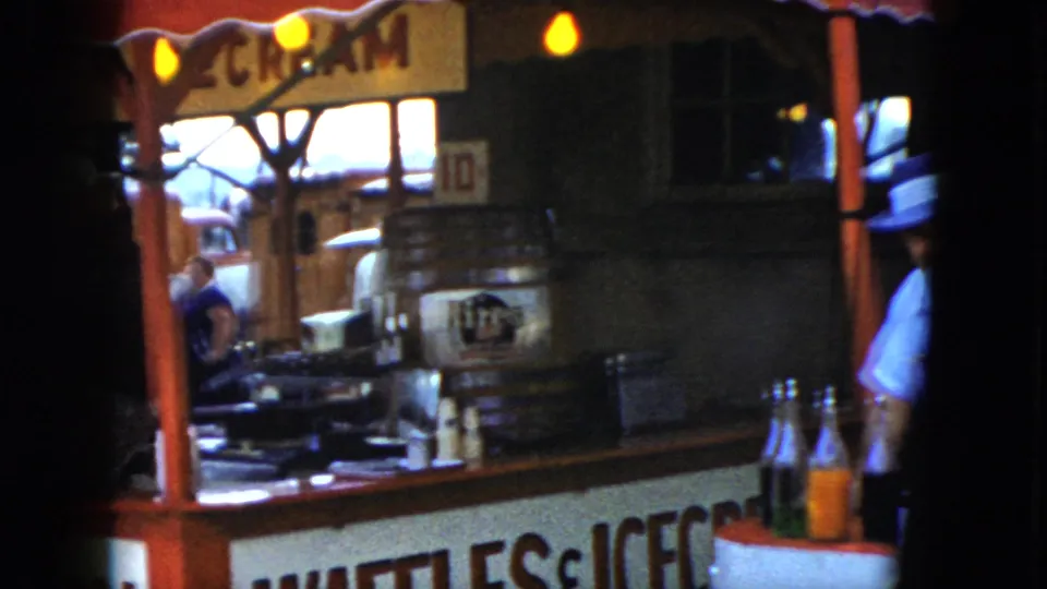 The camera shifts across the 1951 Cleveland booth, showing bottles, partial signage, warm bulbs, counter equipment, and the vendor partly leaving frame.